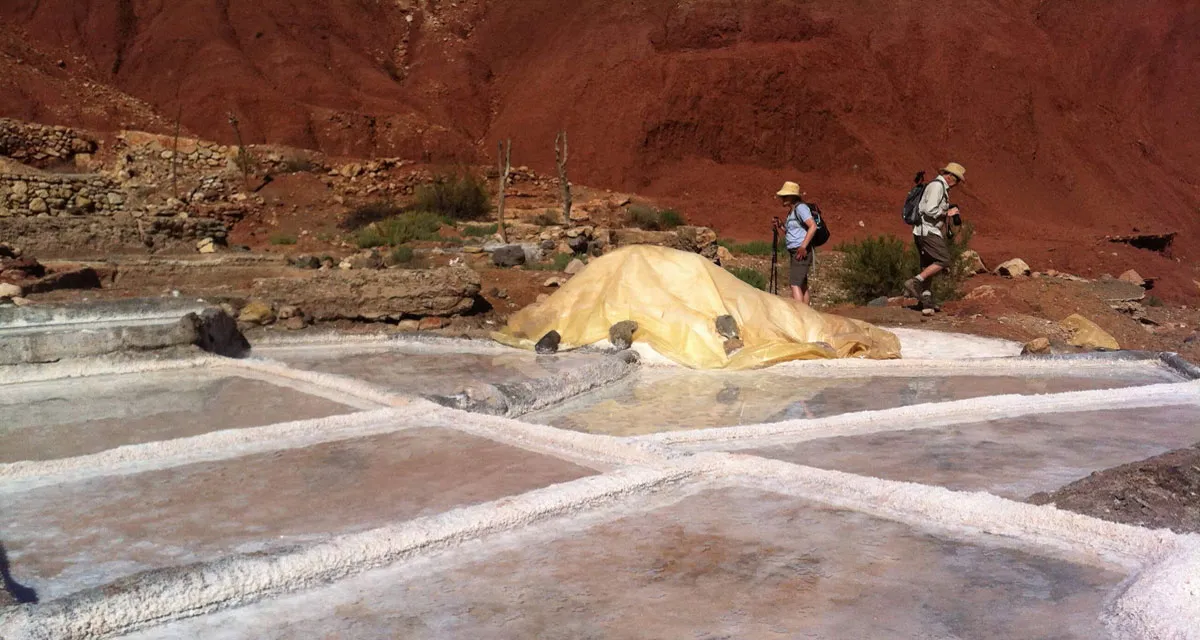 Two trekkers with sun hats and daypacks walking past shallow salt evaporation pans below a red earth hillside near Ouirgane