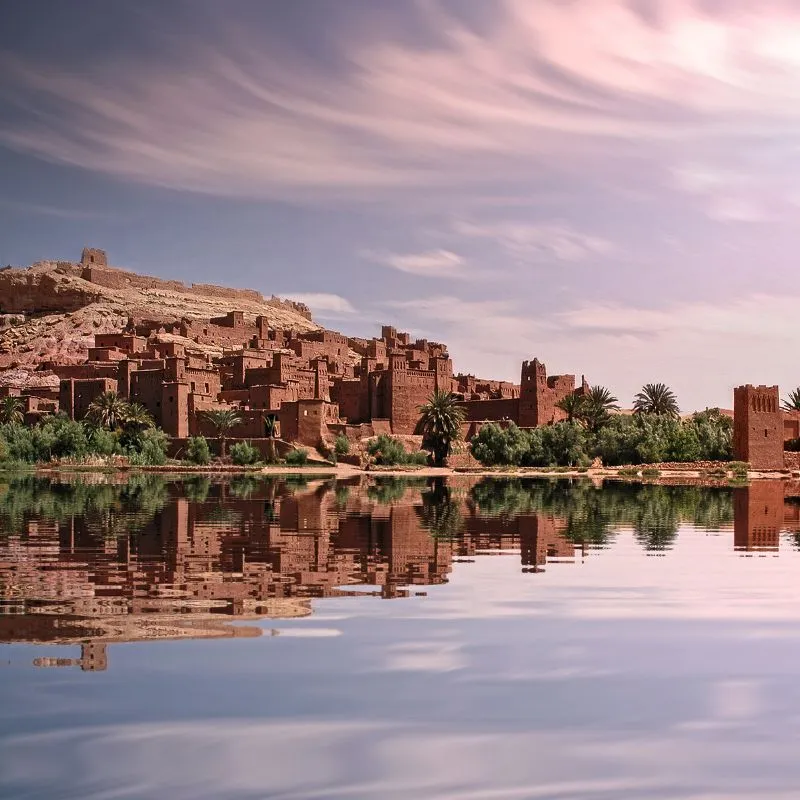 The earthen towers of the Ait Benhaddou ksar reflected in still water under a pink sky